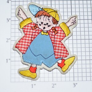 May include: A red and white checkered fabric patch with a cartoon image of a child wearing a blue and yellow outfit, a yellow hat, and a blue cap. The child is smiling and has red hair.