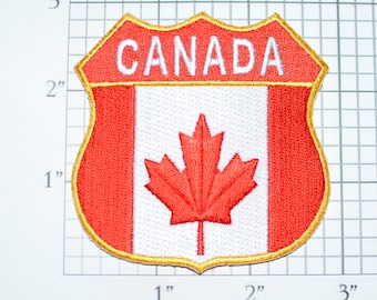May include: A red, white, and gold embroidered patch with the word "CANADA" in white lettering and a red maple leaf on a white background. The patch is shaped like a shield with a gold border.