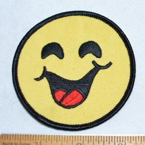 May include: A yellow smiley face with a wide, toothy grin and a red tongue sticking out. The smiley face is embroidered on a black fabric patch.