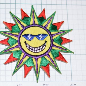 May include: A yellow smiling sun with blue sunglasses and a purple, green, and red starburst design. The sun is wearing sunglasses with lightning bolts on the lenses.