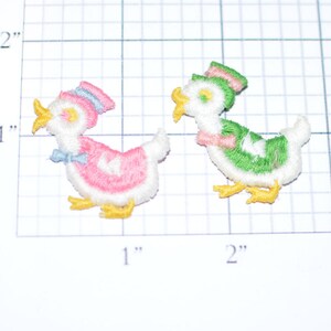 May include: Two embroidered iron-on patches of ducks. One duck is pink with a blue hat and the other is green with a pink hat. Both ducks have yellow beaks and feet.