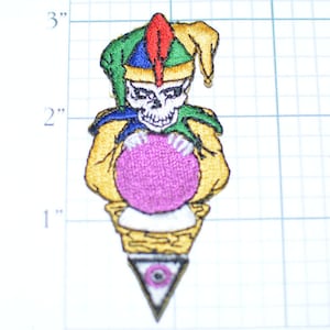May include: Embroidered patch featuring a skull wearing a jester's hat with red, green, blue, and yellow accents. The skull holds a purple orb and is set on a white and gold base with a triangle and eye design. The patch measures approximately 3 inches in height.