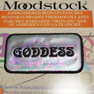 May include: A silver iridescent iron-on patch with black embroidery that reads "GODDESS". The patch is rectangular and has a black border. The patch is for jeans, tops, and jackets.