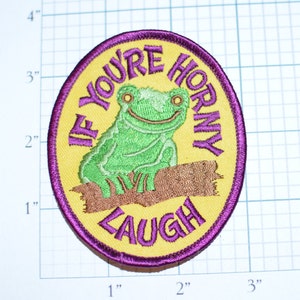 May include: Embroidered patch with a yellow background and a purple border. The patch features a green frog sitting on a brown log, with the text "IF YOU'RE HORNY LAUGH" in purple. The patch measures approximately 3 inches in diameter.
