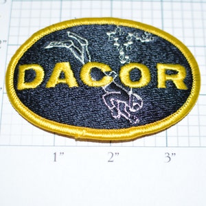 May include: A black and gold embroidered patch with the word "DACOR" in yellow lettering and a white silhouette of a scuba diver in the center.