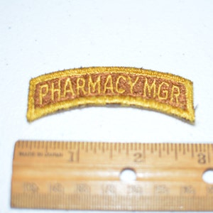 May include: A gold embroidered patch with the text "Pharmacy Mgr" in brown lettering.
