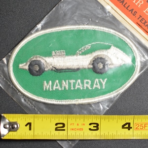 May include: A green oval embroidered patch with a white race car and the text "MANTARAY". The patch is in a plastic bag with a price tag that reads "CAR EMBROIDERED EMBLEMS 69¢ DALLAS, TEXAS."
