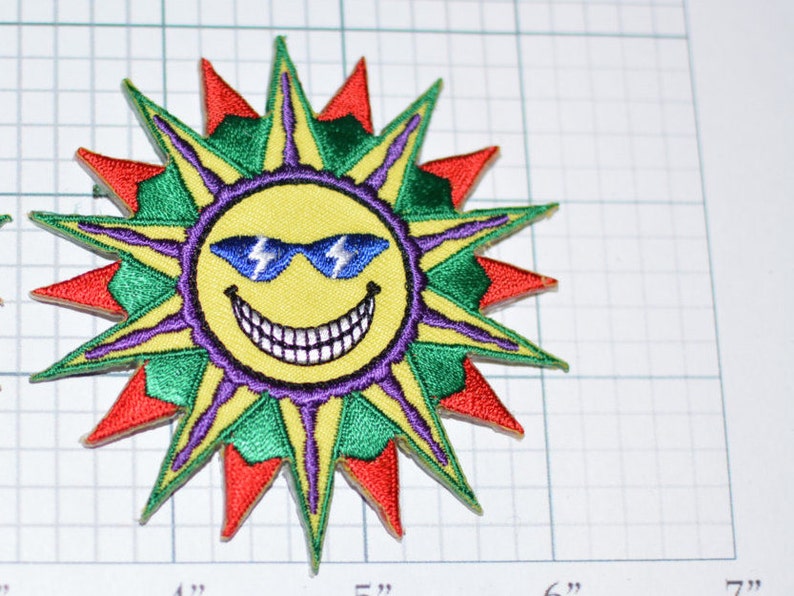 Op de afbeelding: Een gele lachende zon met blauwe zonnebrillen en een stervormig ontwerp in paars, groen en rood. De zon draagt een zonnebril met bliksemschichten op de glazen.