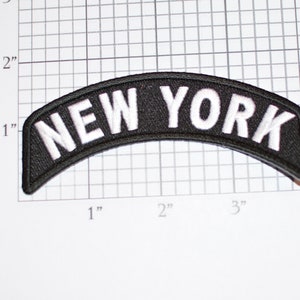 Puede incluir: Un parche negro y arqueado con el texto blanco bordado "NEW YORK". El parche mide aproximadamente 10 cm de ancho y 2,5 cm de alto. El parche est&aacute; sobre un fondo de cuadr&iacute;cula blanca.