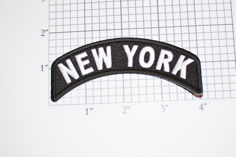 Puede incluir: Un parche negro y arqueado con el texto blanco bordado "NEW YORK". El parche mide aproximadamente 10 cm de ancho y 2,5 cm de alto. El parche est&aacute; sobre un fondo de cuadr&iacute;cula blanca.