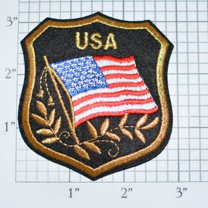 May include: A gold embroidered patch with a black background featuring a stylized American flag with the word "USA" above it. The flag is on a pole with gold embroidered leaves on either side.