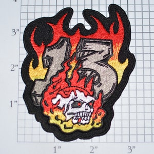 May include: Embroidered iron-on patch featuring the number "13" in gray with flames in red, yellow, and orange. A skull with white teeth and red eyes is in the center of the flames.