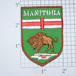 May include: A green and red embroidered patch with the word "MANITOBA" in white lettering. The patch features a white cross on a red background and a brown bison in the center. The patch is approximately 2 inches tall and 1.5 inches wide.