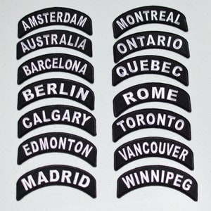 May include: A set of 16 black and white embroidered patches with city and region names. The patches are shaped like an arch and have white lettering on a black background.