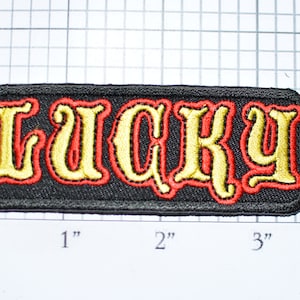 Puede incluir: Un parche de tela negro con la palabra "LUCKY" bordada en oro con un contorno rojo. El parche mide aproximadamente 2,5 cm por 7,5 cm.