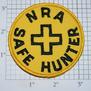 May include: A round, yellow patch with black text and a black cross in the center. The text reads "NRA SAFE HUNTER". The patch is approximately 3 inches in diameter.
