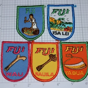 May include: Five embroidered patches with a shield shape and a blue, green, yellow, or orange background. Each patch features the word "Fiji" in white lettering and a different image. The images include a person making a drum, a beach scene, a wooden club, a wooden stick, and a necklace.