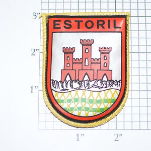 May include: A red, white, and green embroidered patch with the word "ESTORIL" in black lettering. The patch features a red castle with three towers and a green and white checkered pattern at the bottom.