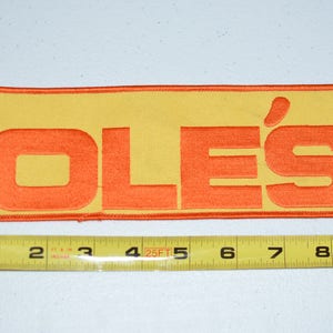 May include: A yellow and orange embroidered patch with the word "OLE'S" in orange lettering.