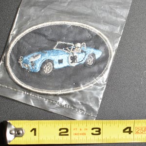 May include: A blue and white embroidered patch of a vintage sports car on a black background. The car has a driver in the seat and the number 9 on the side.