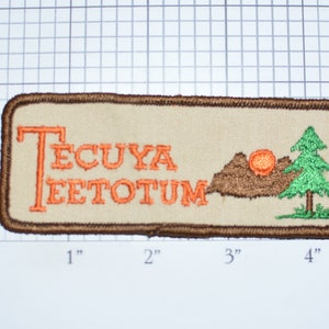 May include: A brown rectangular patch with a beige background. The patch has the text "TECUYA EETOTUM" in orange embroidery. There is a brown mountain with an orange sun and a green pine tree on the right side of the patch.