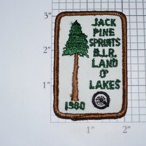 May include: A white embroidered patch with a brown border. It features a green pine tree, the text "Jack Pine Sprints BLR. Land O' Lakes", and the year "1980".