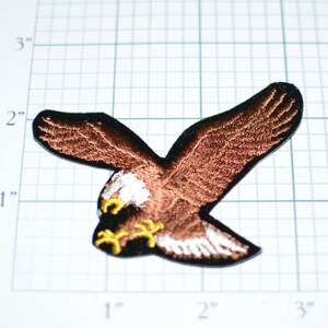 May include: Brown embroidered eagle patch with white and yellow accents. The eagle is in flight with its wings spread wide. The patch is approximately 2 inches wide.