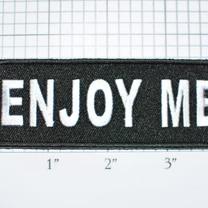 May include: Black rectangular patch with the words "ENJOY ME" embroidered in white block letters. The patch is approximately 4 inches long and 1 inch high.