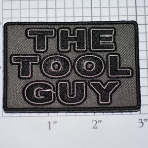 May include: A gray embroidered patch with white outlines. The patch reads "THE TOOL GUY" in black block letters.