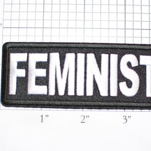 May include: A rectangular black patch with the word "FEMINIST" embroidered in white, bold capital letters. The patch measures approximately 4 inches long and 1 inch high.