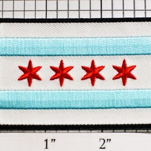 May include: A white patch with a blue and white striped design and four red stars. The patch is surrounded by a black border. The patch is approximately 2 inches wide and 1 inch tall.