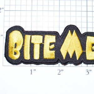 May include: A black and gold embroidered patch with the text "BITE ME" in yellow lettering.
