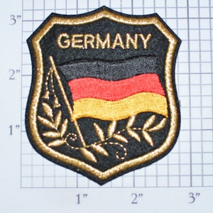 May include: A black and gold embroidered patch with the German flag and the word "GERMANY" in gold lettering. The patch is shaped like a shield and has a gold border. It is approximately 3 inches wide and 3 inches tall.