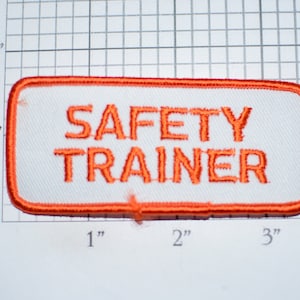 May include: Orange embroidered patch with white background and the text "SAFETY TRAINER" in orange.