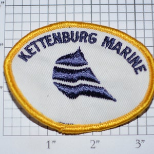 May include: A white oval patch with a yellow border. The patch has the text "KETTENBURG MARINE" in blue embroidery around the edge. A blue and white stylized ship's wheel is in the center.