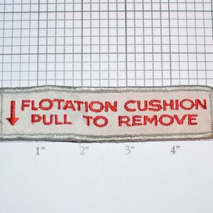 May include: A white rectangular patch with red embroidered text that reads "FLOTATION CUSHION PULL TO REMOVE". A red arrow pointing down is to the left of the text.