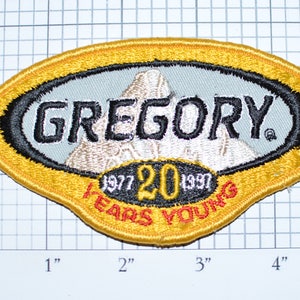 May include: A yellow, oval-shaped patch with a black border and white stitching. The patch features a white mountain range with the text "GREGORY" in black lettering. Below the mountain range is a black oval with the text "1977 20 1997 YEARS YOUNG" in white and red lettering.