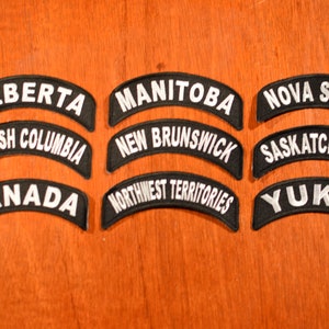 May include: Nine black and white embroidered patches with the names of Canadian provinces and territories. The patches are arranged in three rows of three. The patches are on a wooden surface.
