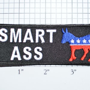 May include: Black rectangular patch with the words "SMART ASS" in white block letters. A blue donkey with white stars and red accents is on the right side. The patch measures approximately 4 inches wide and 2 inches high.