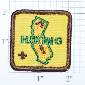 May include: A yellow embroidered patch with a green outline of a hiking boot and the word "HIKING" in green letters. A brown scout symbol is in the lower left corner.