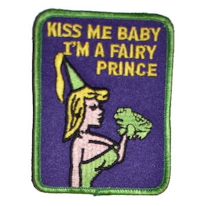 May include: A purple embroidered patch with a green border. The patch features a cartoon image of a woman wearing a green dress and a green hat. She is holding a green frog in her right hand. The text "Kiss Me Baby I'm A Fairy Prince" is written in yellow letters on the patch.