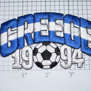 May include: Embroidered patch featuring the word "GREECE" in blue with white stripes, a white soccer ball with black panels, and the numbers "1994" in white.
