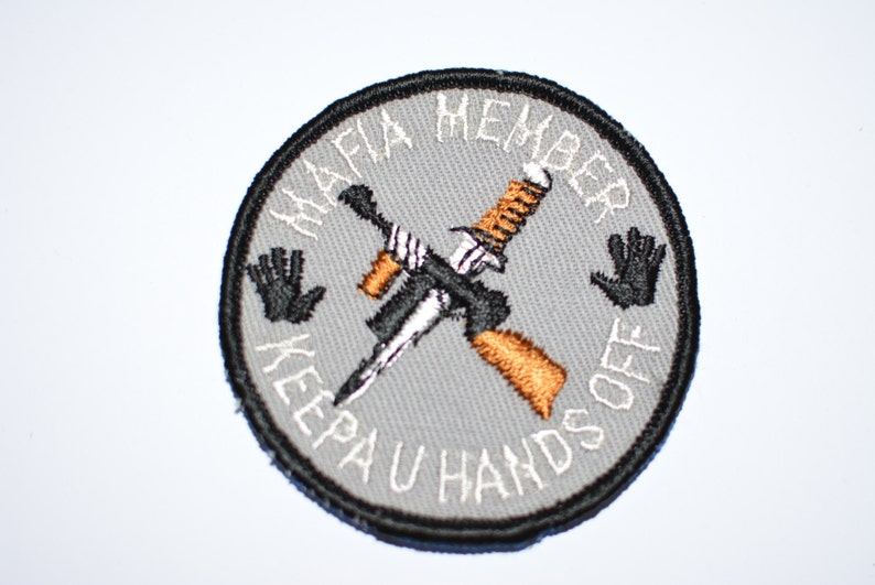 May include: A gray circular patch with black stitching around the edge. The patch has white text that reads "Mafia Member Keep Your Hands Off" with a crossed knife and gun in the center. The knife is silver and the gun is brown.