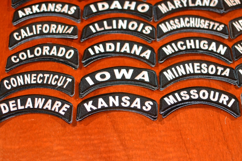 Puede incluir: Primer plano de varios parches bordados en blanco y negro. Cada parche es curvo y presenta el nombre de un estado en letras may&uacute;sculas blancas. Los estados incluyen Arkansas, California, Colorado, Connecticut, Delaware, Iowa y Kansas.