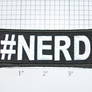 May include: Black fabric patch with white embroidered text that reads "#NERD". The patch is approximately 3 inches wide and 1 inch tall.