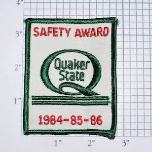 May include: A white patch with green and red embroidery. The patch reads "SAFETY AWARD" in red lettering, "Quaker State" in green lettering, and "1984-85-86" in red lettering.