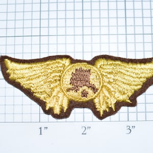 May include: A brown and gold embroidered patch with a winged design. The wings are gold and the center of the patch is brown with a stylized tree design.