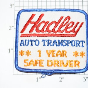 May include: A white embroidered patch with blue edging. The patch features the text "Hadley Auto Transport" in blue lettering, "1 Year" in orange lettering, and "Safe Driver" in orange lettering. The word "Hadley" is in red lettering.