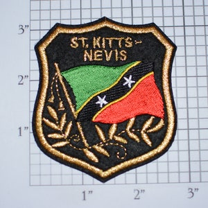 May include: A black and gold embroidered patch featuring the flag of Saint Kitts and Nevis. The flag is green, black, and red with two white five-pointed stars. The patch is shaped like a shield and has the words "ST. KITTS-NEVIS" embroidered in gold.