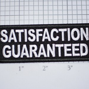 May include: A rectangular black patch with the words "SATISFACTION GUARANTEED" embroidered in white. The patch measures approximately 4 inches wide and 2 inches tall.